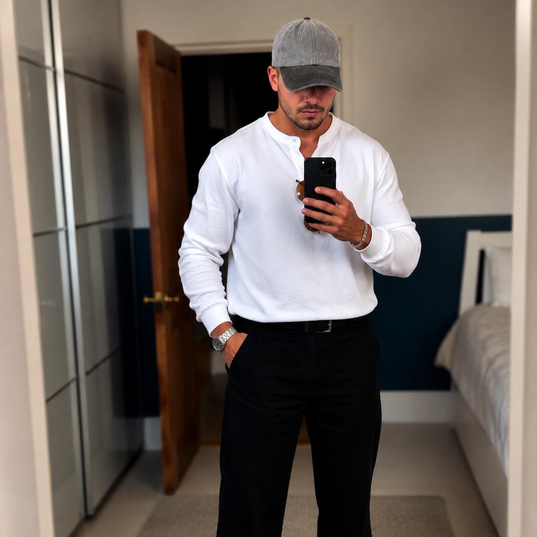 A man takes a mirror selfie in a bedroom, wearing a white long-sleeve henley shirt, black trousers, black and white sneakers, and a gray baseball cap.
