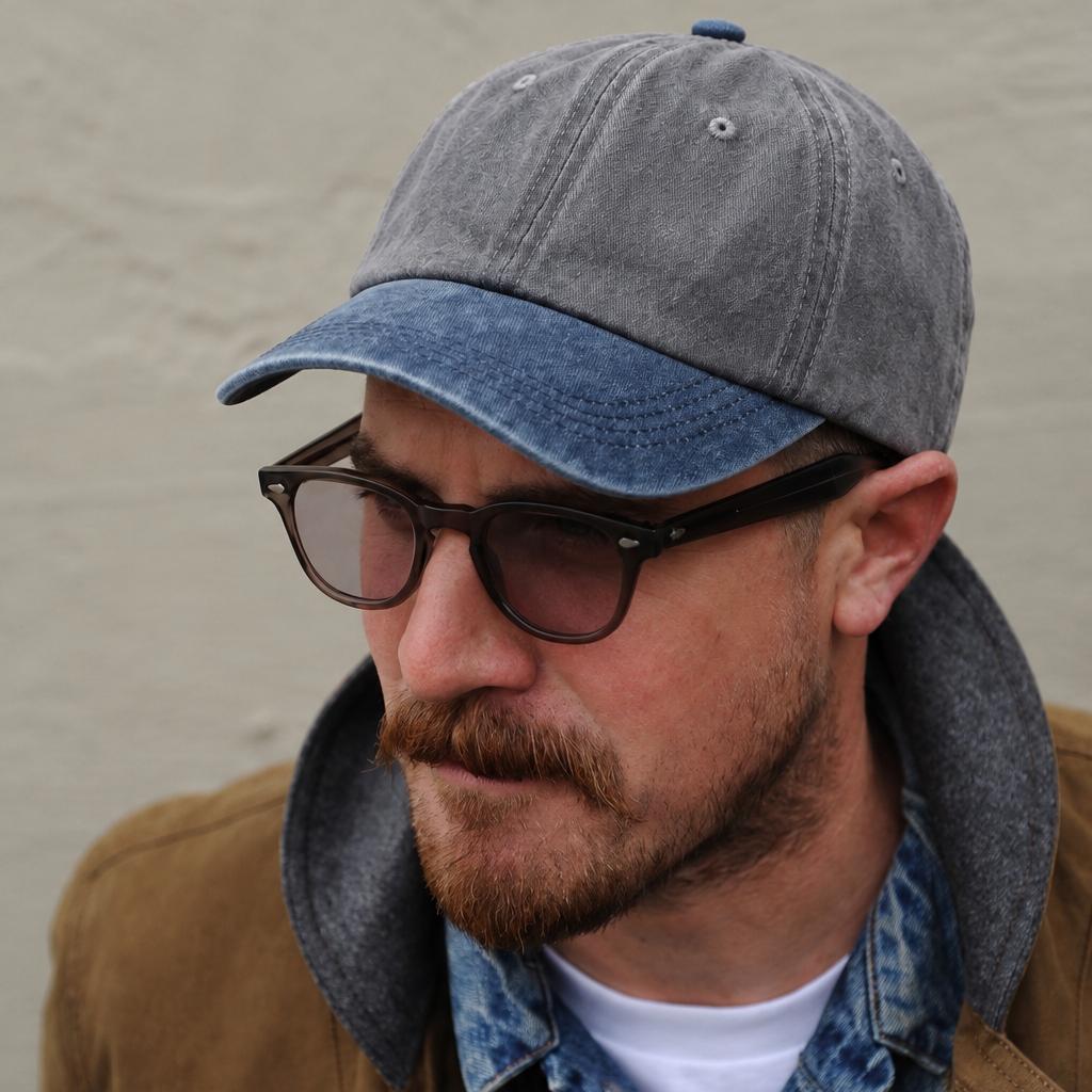 A man wearing a gray and blue distressed baseball cap, black sunglasses, a brown jacket, and a denim shirt, photographed against a textured beige wall.
