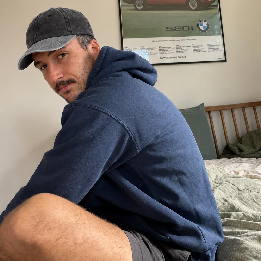 A man wearing a navy blue hoodie and a dark baseball cap sits on a bed in a bedroom, looking back over his shoulder. A framed BMW S2000 car poster is hanging on the wall behind him.