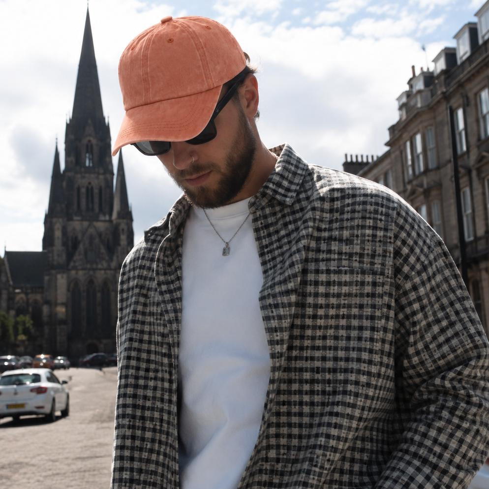 A man wearing a black and white checkered flannel shirt over a white t-shirt, light blue jeans, and an orange baseball cap, standing on a cobblestone street with a historic cathedral in the background.