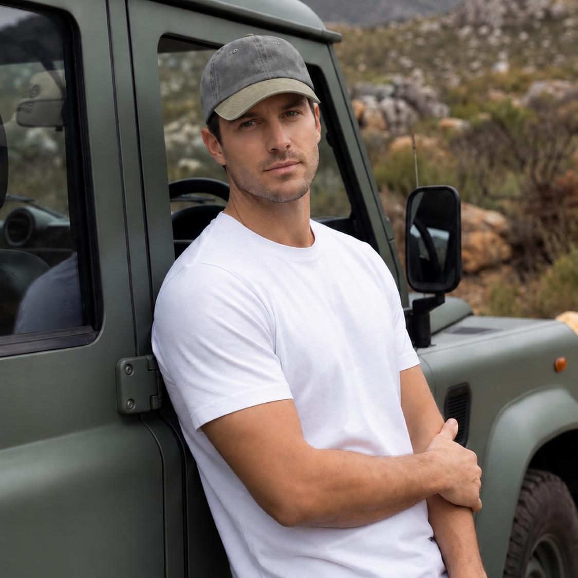 A man wearing a white crewneck t-shirt and a gray baseball cap leans against an olive green off-road vehicle, with a rocky mountain landscape in the background.