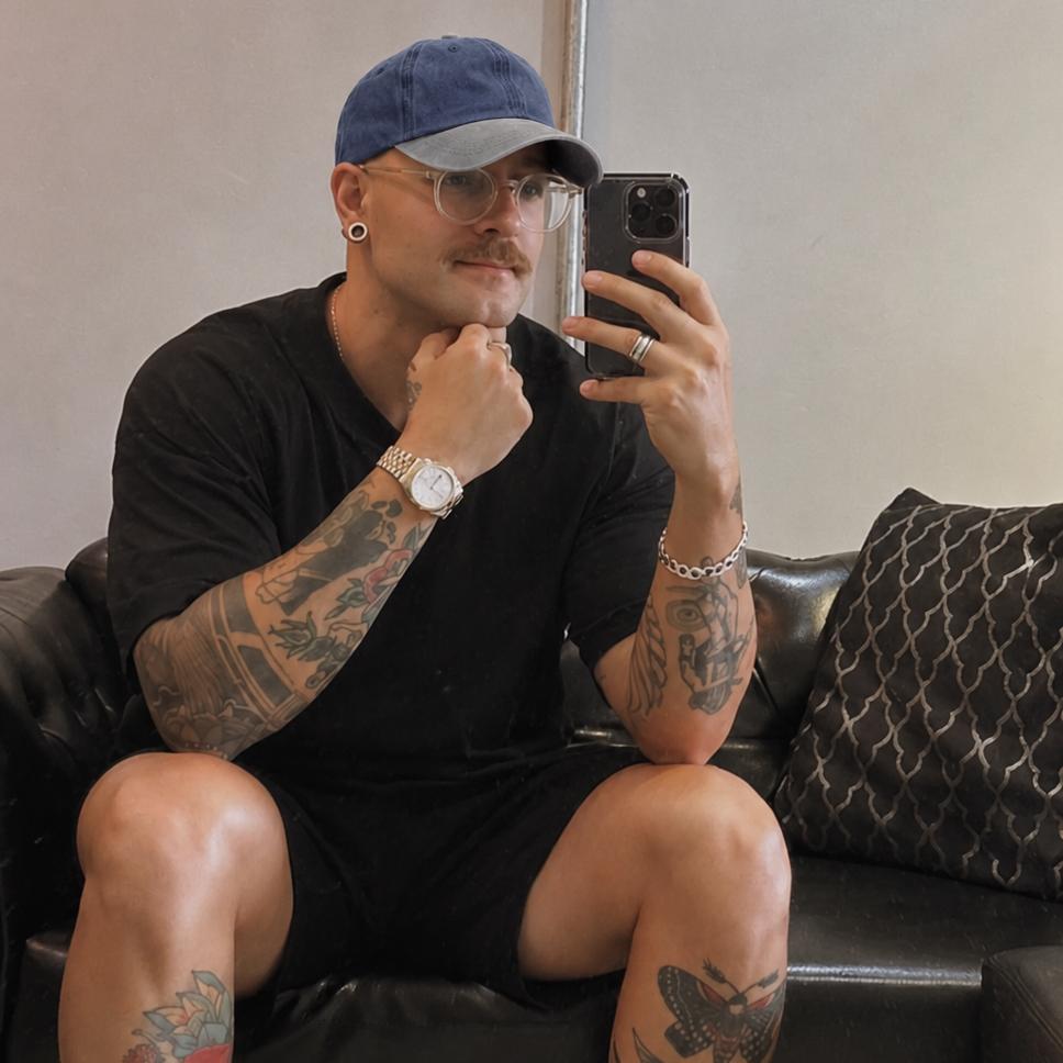 A man with tattoos takes a mirror selfie while wearing a black t-shirt, black shorts, a blue baseball cap, and white sneakers, sitting on a black leather couch.