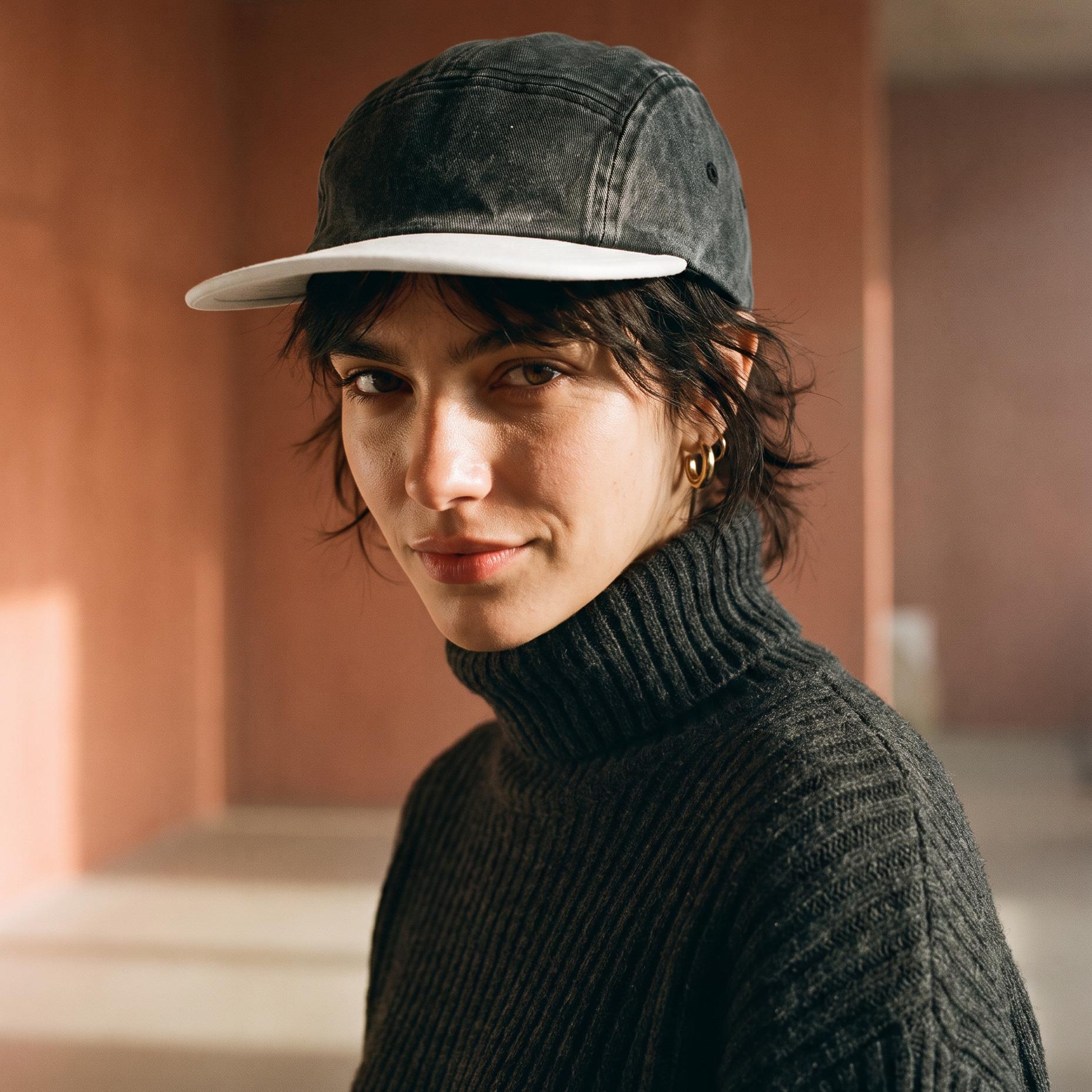 A person wearing a washed black baseball cap with a white brim and a dark ribbed turtleneck sweater, photographed in a warm-toned indoor space with terracotta walls.