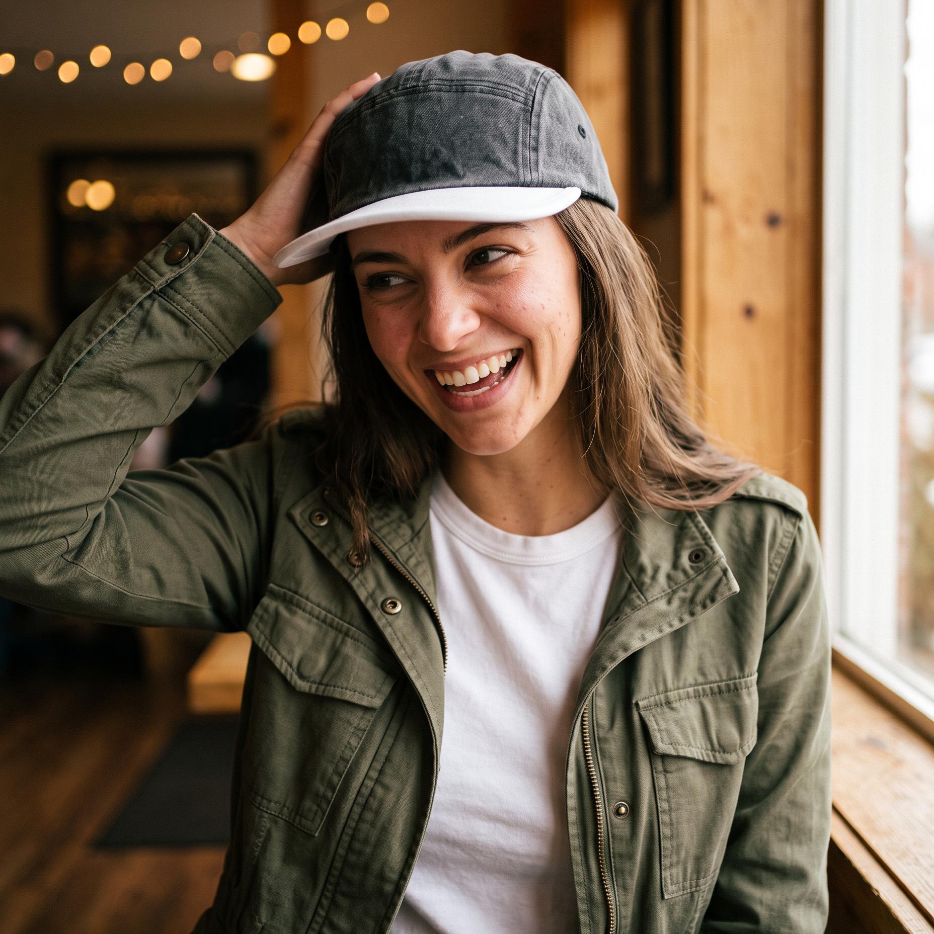 A woman smiling in a cozy indoor setting, wearing an olive green utility jacket over a white t-shirt and a dark gray and white baseball cap.