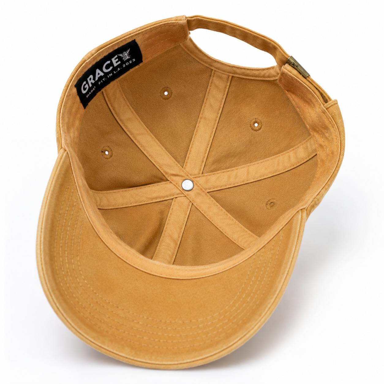 A tan canvas baseball cap shown from an overhead angle, displaying the interior crown, adjustable strap, ventilation eyelets, and a black interior label that reads 'GRACE'.