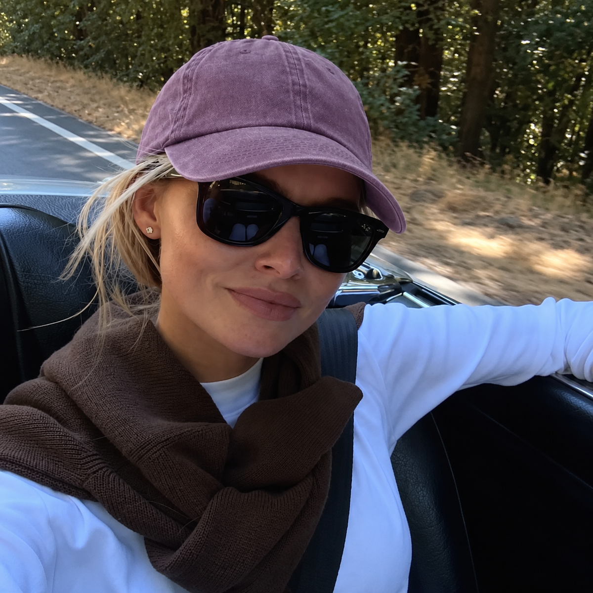 A woman wearing a dusty mauve baseball cap, black sunglasses, a brown knit scarf, and a white long-sleeve shirt sits in a convertible car on a road with trees in the background.
