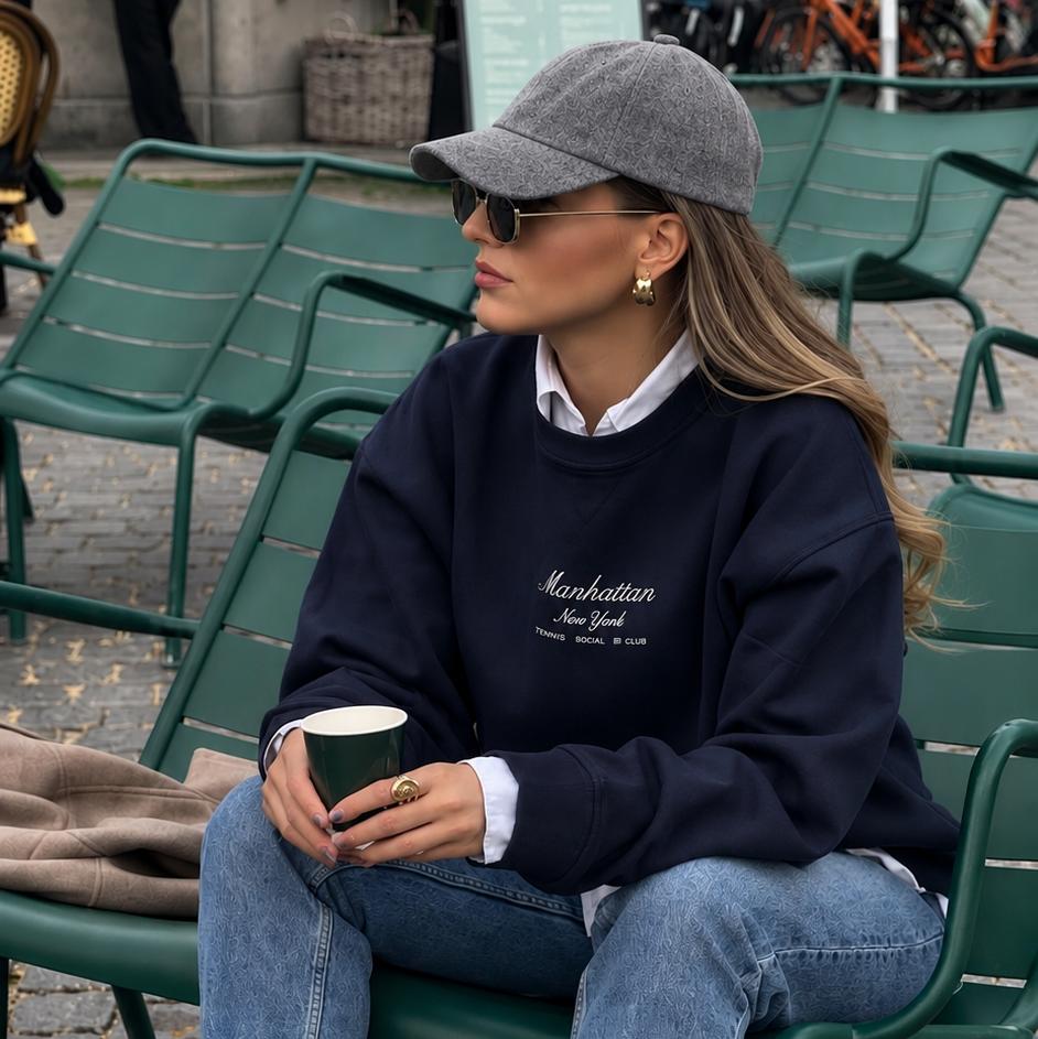 A woman sits on a green metal cafe chair wearing a navy blue sweatshirt with 'Manhattan New York' printed on the chest, light blue jeans, white crew socks, and cream sneakers with black stripes. She is holding a paper coffee cup in an outdoor cafe setting.