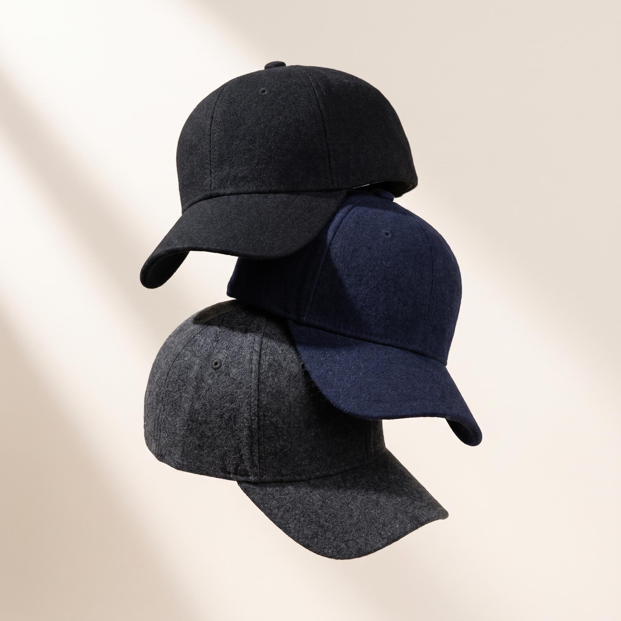 Three baseball caps stacked together on a warm off-white background with soft diagonal shadows. The top cap is black, the middle cap is navy blue, and the bottom cap is charcoal gray.